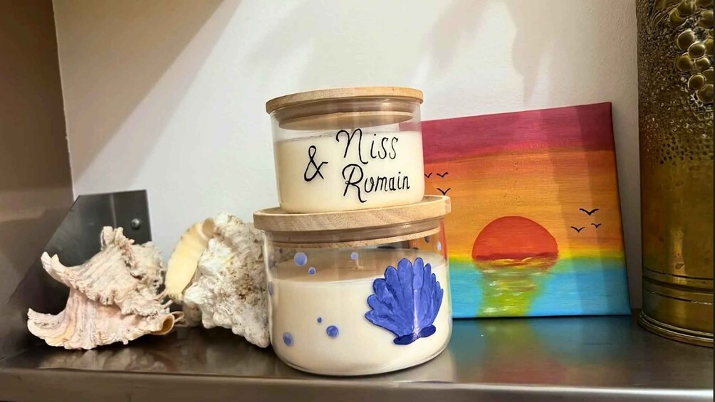 A decorative jar with 'Nissa & Romain' written on it, placed on a shelf with seashells and a sunset painting. 4