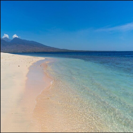 A beach with clear turquoise waters, a sandy shore, and distant mountains under a clear sky. 1