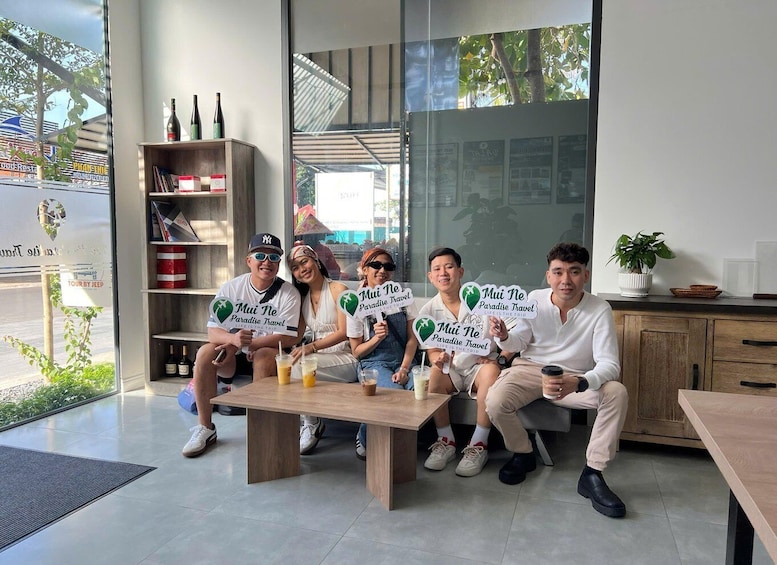 A group of people sitting at a wooden table in a modern indoor setting with a glass wall, a shelf with bottles, and a sign for 'TOUR BY JEEP'. 5