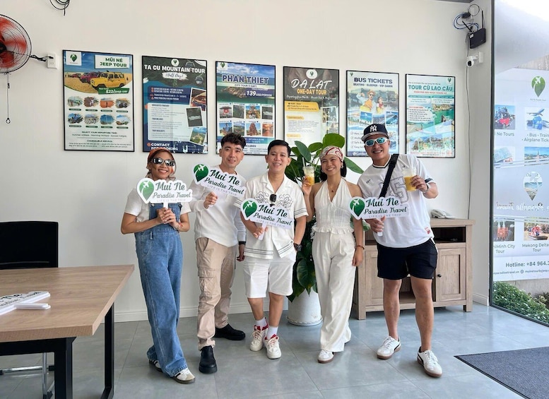 A group of people holding signs with 'MUI NE Paradise Travel' branding, standing in an indoor setting with travel posters on the wall. 4