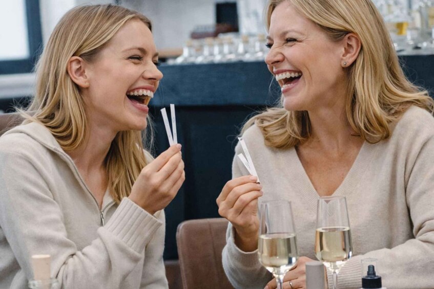 Two people sitting at a table with glasses of wine and a bottle of perfume. 1
