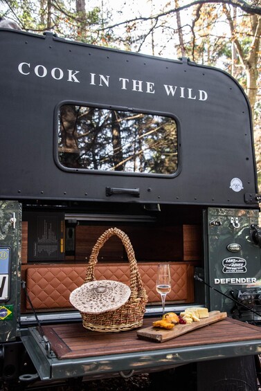 A camper with 'COOK IN THE WILD' written on the side, equipped with a wicker basket, a glass of wine, and a wooden board with food. 1
