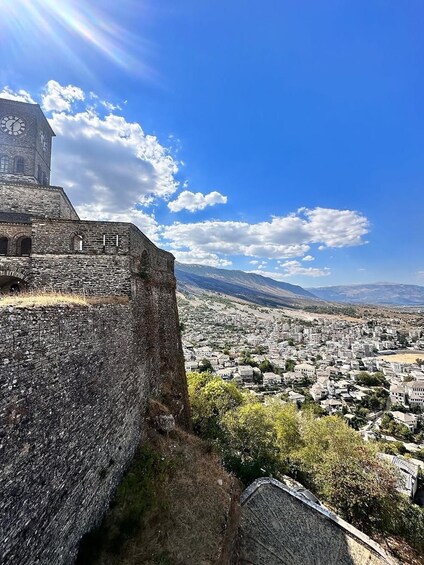 A historic stone wall with a clock tower, overlooking a town with mountains in the distance. 4