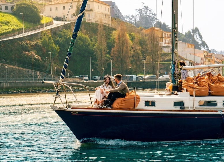 A sailboat with two people on deck, a calm water body, and buildings in the background. 1