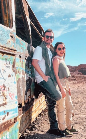 A couple standing by a rusted, graffiti-covered vehicle in a desert landscape. 5