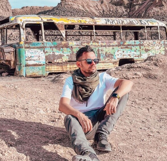 A man sitting on rocky ground with an abandoned bus in the background. 2