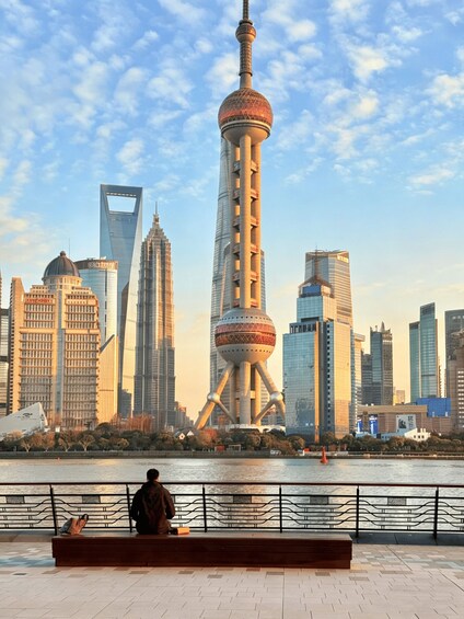 A person sitting on a bench overlooking a river with a city skyline in the background, including a distinctive tower with a spherical observation deck. 2