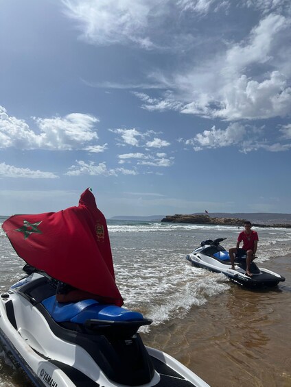 Two jet skis on a beach with a flag on one and a person sitting on the other. 3