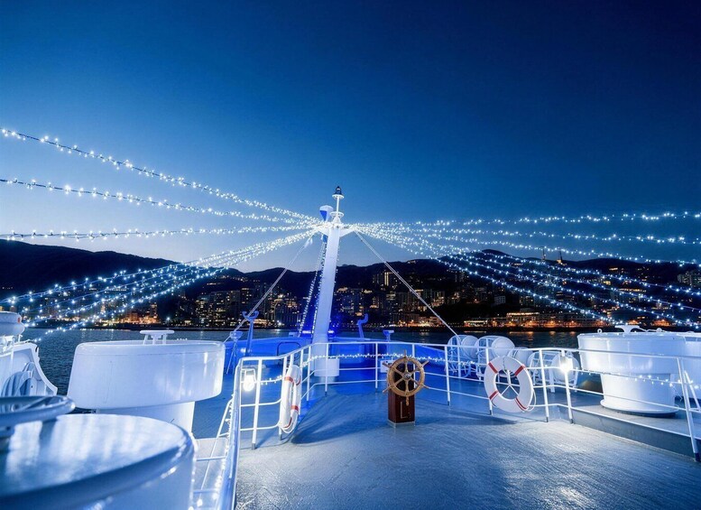 A boat deck with white tables, a lifebuoy, and a steering wheel, with string lights overhead and a cityscape in the background. 3