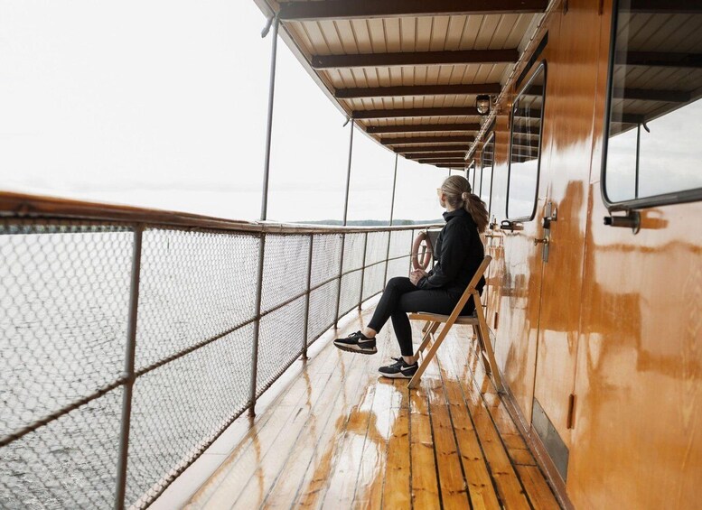 A person sitting on a wooden bench on a boat deck, with a metal railing and a view of the water. 3