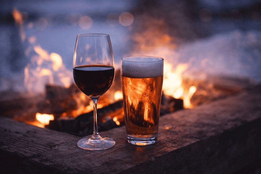 A glass of red wine and a glass of beer by a campfire. 4