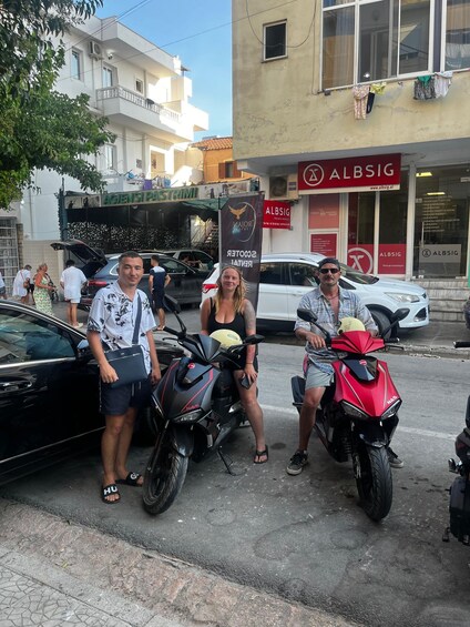 A street scene with parked motorcycles, a building with 'ALBSIG' signage, and people standing nearby. 5