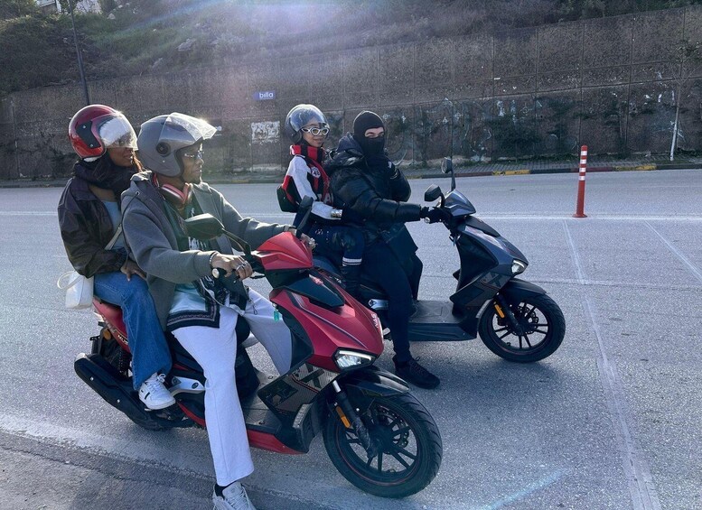 Three individuals on a red motorcycle and a black scooter, with a concrete barrier and a red and white striped pole in the background. 1