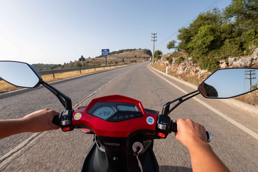 A red motorcycle with a digital speedometer, a hand on the handlebar, and a road with a blue sign in the distance. 4