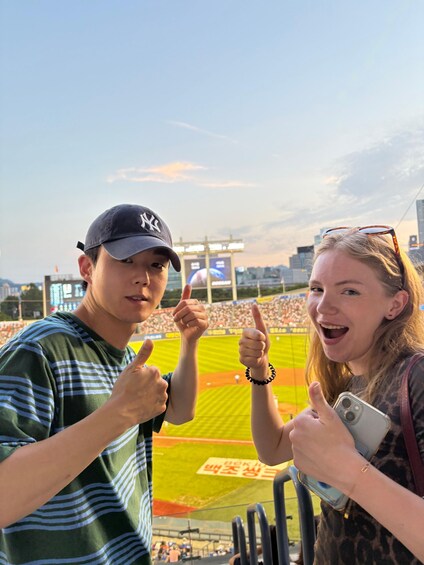 Two people giving thumbs up at a baseball stadium with a city skyline in the background. 2