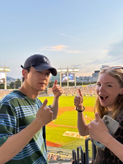 Two people giving thumbs up at a baseball stadium with a large screen and a crowd in the background. 1