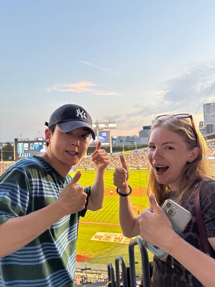 Two people giving thumbs up at a baseball stadium with a city skyline in the background. 5