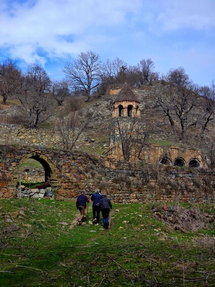 A stone structure with arches, a small tower, and a group of people hiking in a grassy area. 5