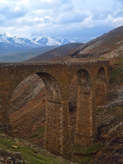 A stone viaduct with multiple arches spanning a valley, surrounded by mountains and a cloudy sky. 1