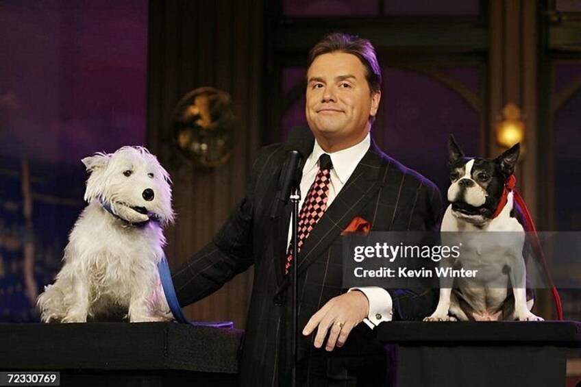 A man in a suit with a microphone, standing next to two dogs. 3