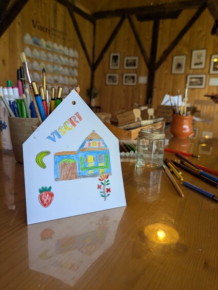 A drawing of a house with a strawberry and a flower, surrounded by art supplies on a wooden table. 3