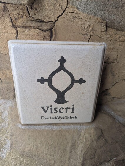 A square ceramic tile with a black emblem and the text 'Viscri Deutsch-Weißkirch' against a stone wall. 2