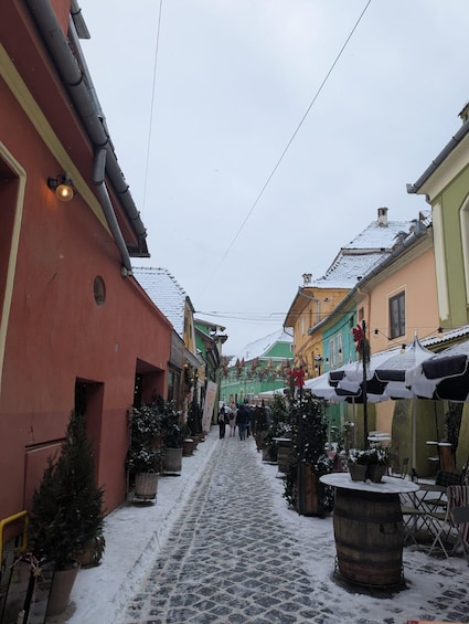 A snow-covered cobblestone street lined with colorful buildings and outdoor seating areas. 5