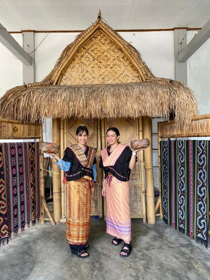 Two individuals in traditional attire standing in front of a thatched-roof structure with patterned textiles on the sides. 4