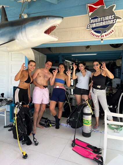 A group of people in scuba diving gear posing in front of a shark sculpture and a diving center sign. 3