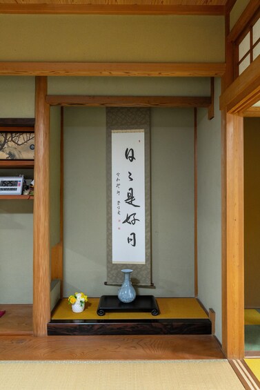 A traditional Japanese room with tatami flooring, a wooden shelf, a vase with flowers, and a calligraphy scroll. 3