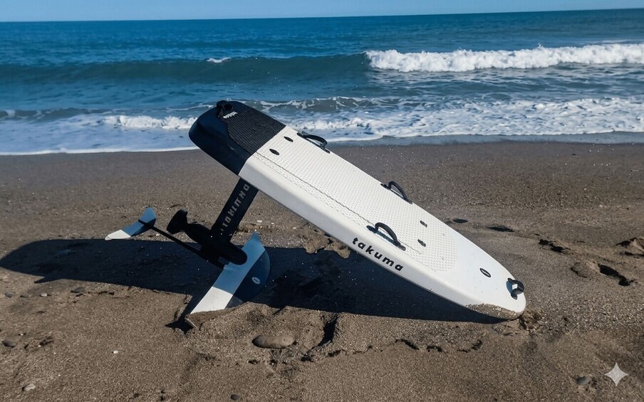 A white paddleboard with 'takuma' written on it, leaning against a blue stand on a sandy beach with waves in the background. 2