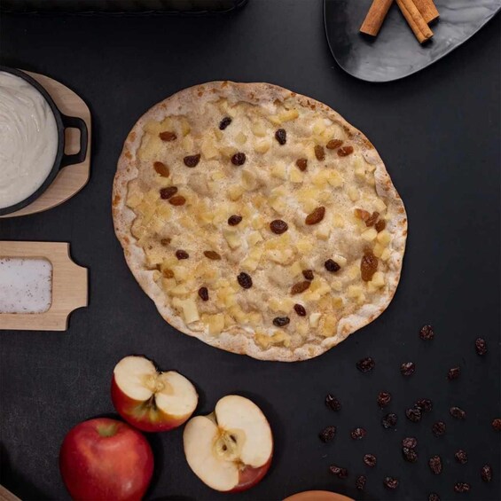 A flatbread with raisins and diced fruit, surrounded by apples and cinnamon sticks on a dark surface. 5