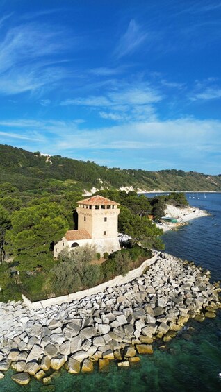 A coastal landscape with a tower, rocky shoreline, clear water, and a forested hill. 5