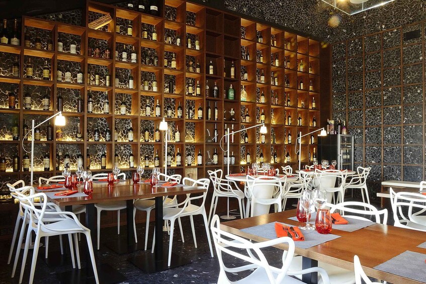A restaurant with white chairs and tables, a wall of shelves with bottles, and a marble wall. 3