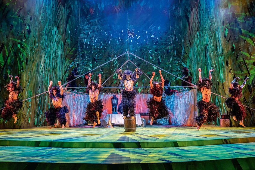 A stage with performers in feathered costumes, a central barrel, and a backdrop of vertical foliage. 5