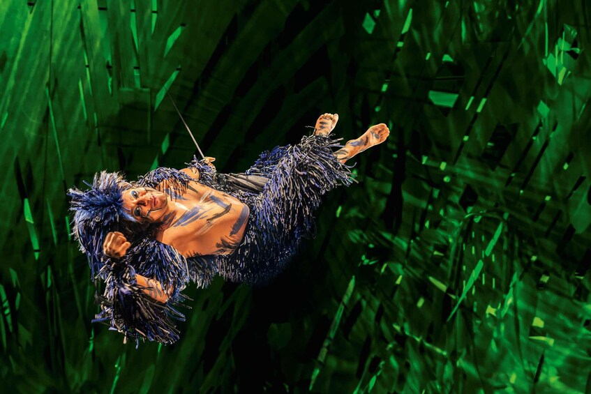 A person in a blue, feathered costume suspended in mid-air against a green, leafy background. 2