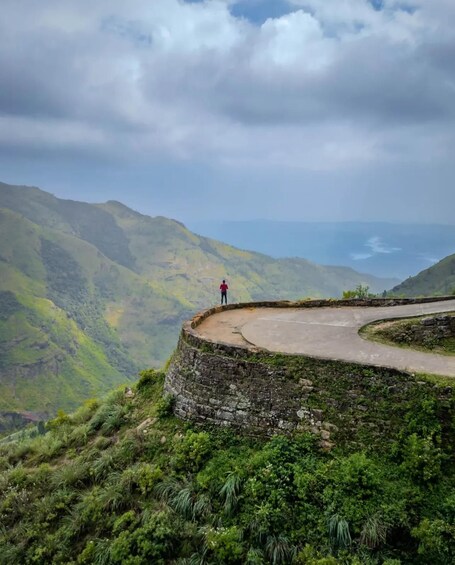 A person standing on a stone platform overlooking a winding road and lush green mountains. 1