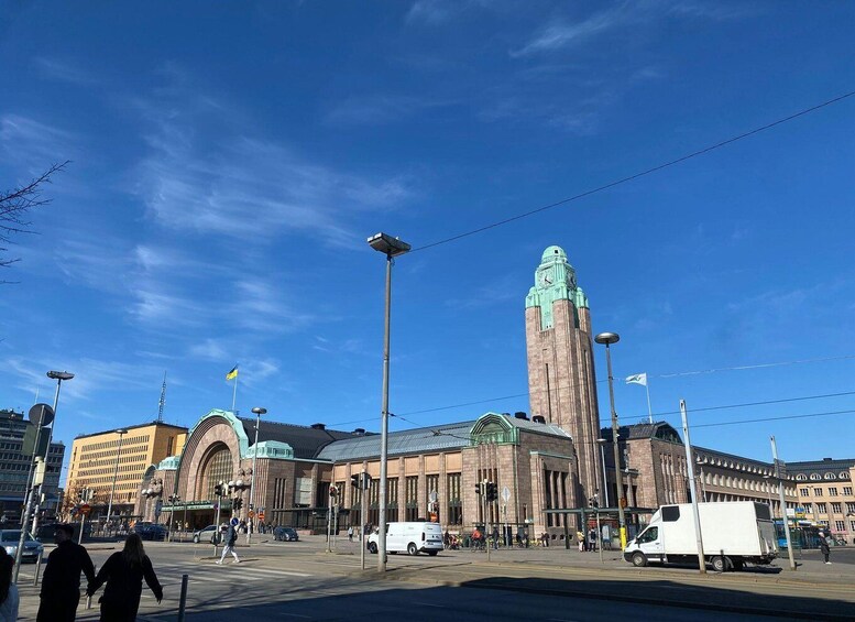 A city street with a prominent clock tower, a tram stop, and a variety of buildings. 3