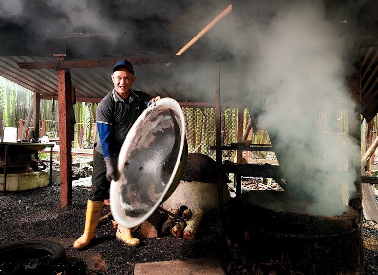A person holding a large, round, metal object in an outdoor setting with smoke rising from a structure. 5