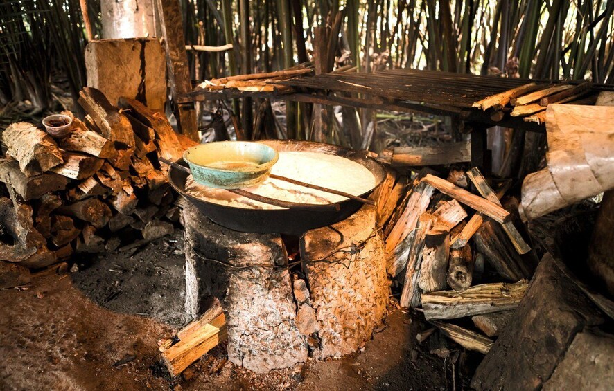 A rustic outdoor cooking setup with a pot on a makeshift stove, surrounded by wood and a woven bamboo structure. 2