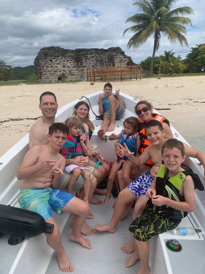 A group of people on a boat on a sandy beach with palm trees and a stone structure in the background. 3