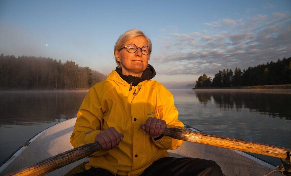 A person in a yellow jacket rowing a boat on a calm lake with a forested shoreline. 1