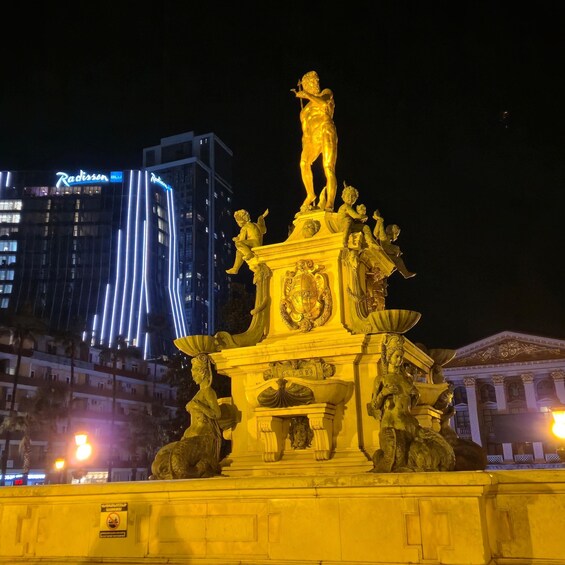 A golden statue of a man playing a lyre, surrounded by smaller figures, with a building in the background. 5