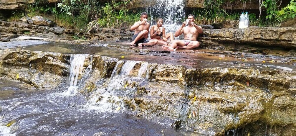 A waterfall, three people sitting on rocks, and a water bottle on a ledge. 3