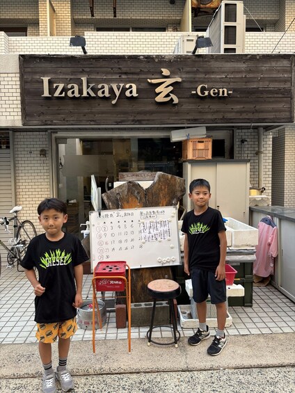 Two children standing in front of a wooden signboard with Japanese characters, a whiteboard with numbers, and a bicycle parked to the left. 5
