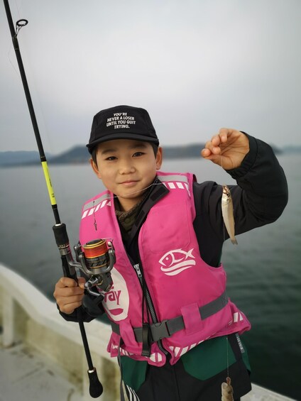 A person holding a fishing rod and a fish, wearing a pink life vest and a cap with motivational text. 1