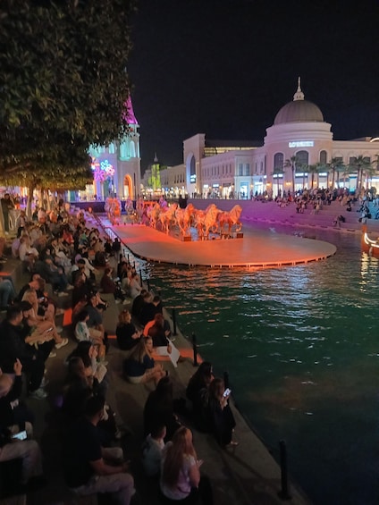 A nighttime outdoor event with a water fountain and illuminated horses, surrounded by spectators. 1