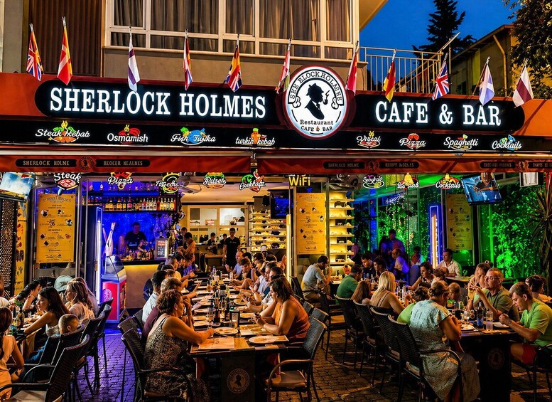 A busy outdoor dining area at Sherlock Holmes Cafe & Bar with various food and drink options. 1