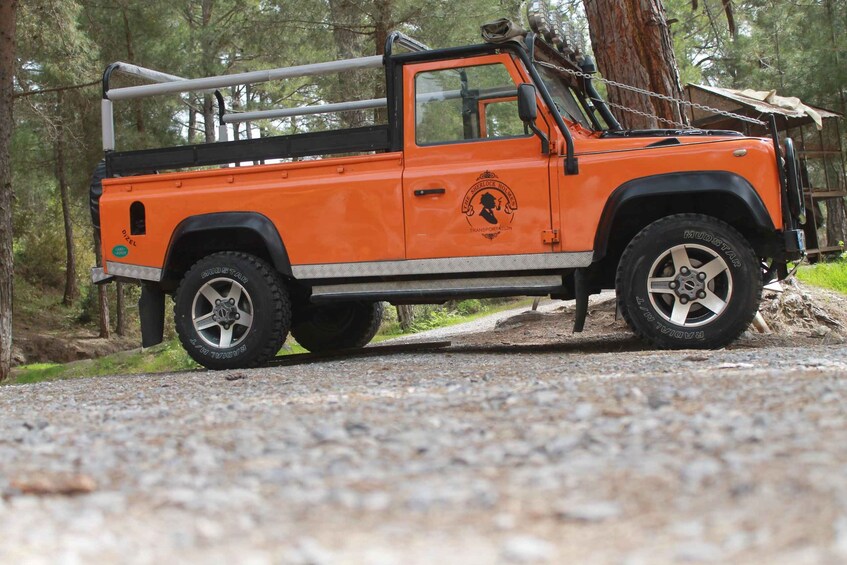 An orange utility vehicle with a ladder and a logo on the side, parked on a gravel path in a wooded area. 2