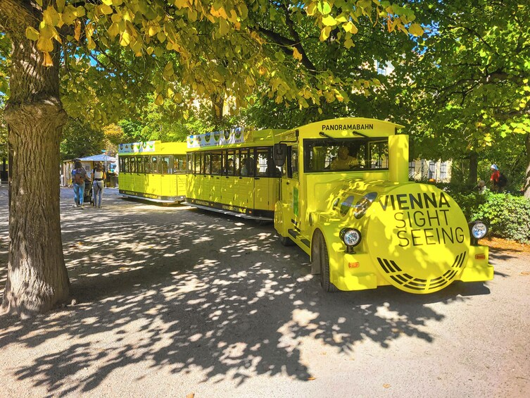 Ein gelber Straßenbahn mit der Aufschrift "Vienna Sightseeing", geparkt unter einem Baum. 1